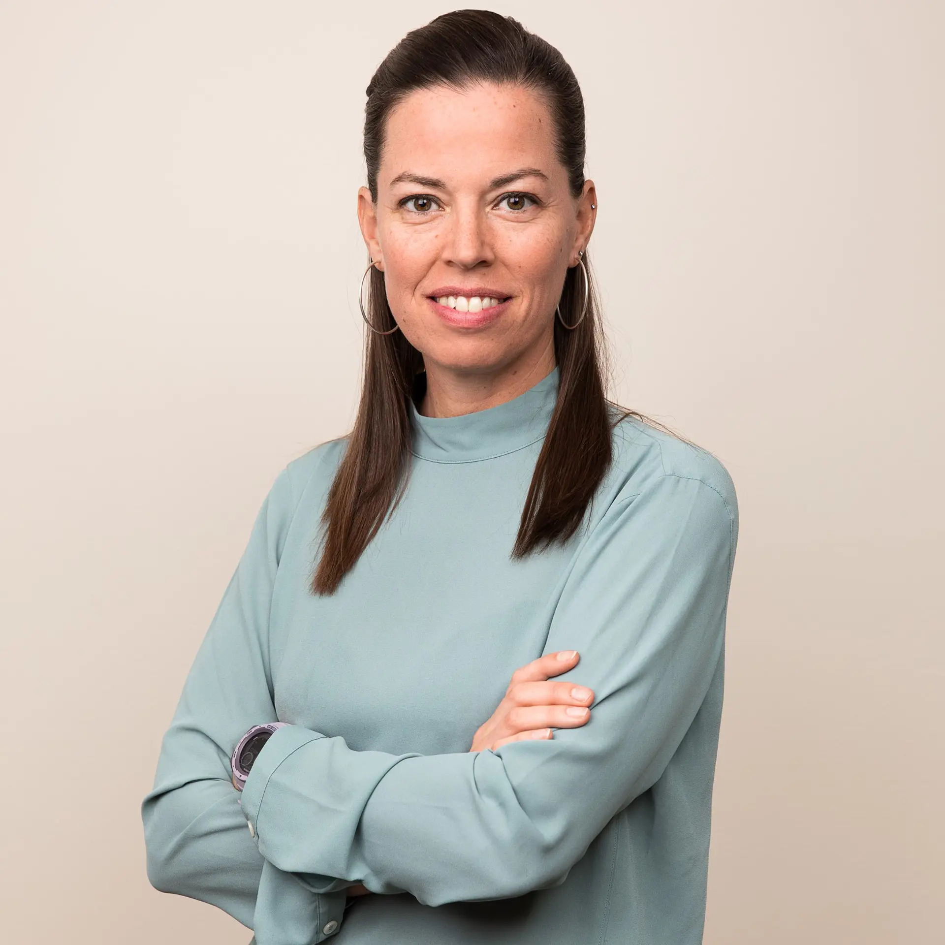 Kvinna med ljus blus, långt mörkt hår står med armarna i kors och tittar in i kameran och ler. 
