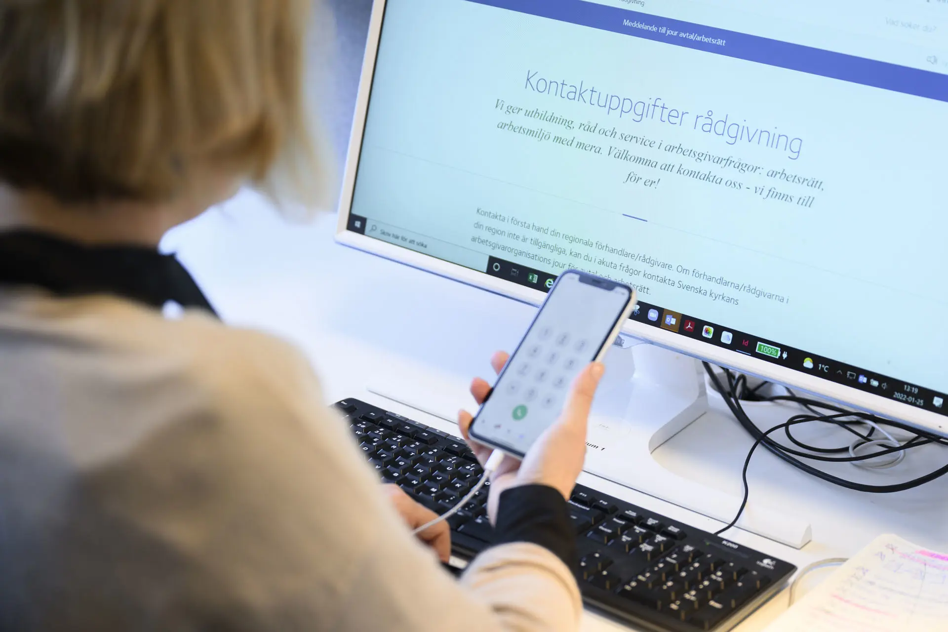 kvinna sitter framför datorskärm med en mobiltelefon i ena handen. 