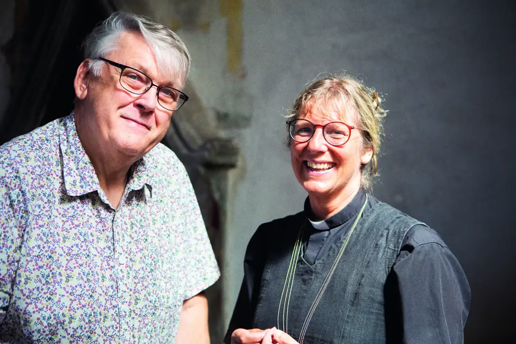 till vänster man i skorta och glasögeon. han lutr sig en aning mot kvinna till höger, kvinna som ler, klädd i prästskjorta och glasögon.