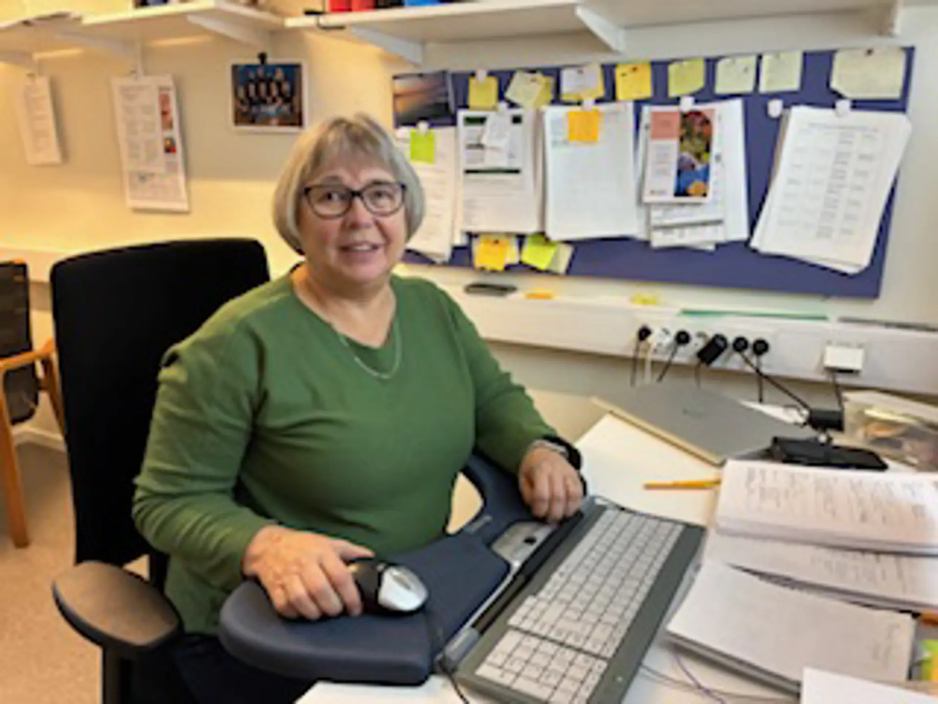 kvinna med grön tröja, pagefrisyr och glasögon sitter vid ett skrivbord, tittar in i kameran och ler svagt. 
