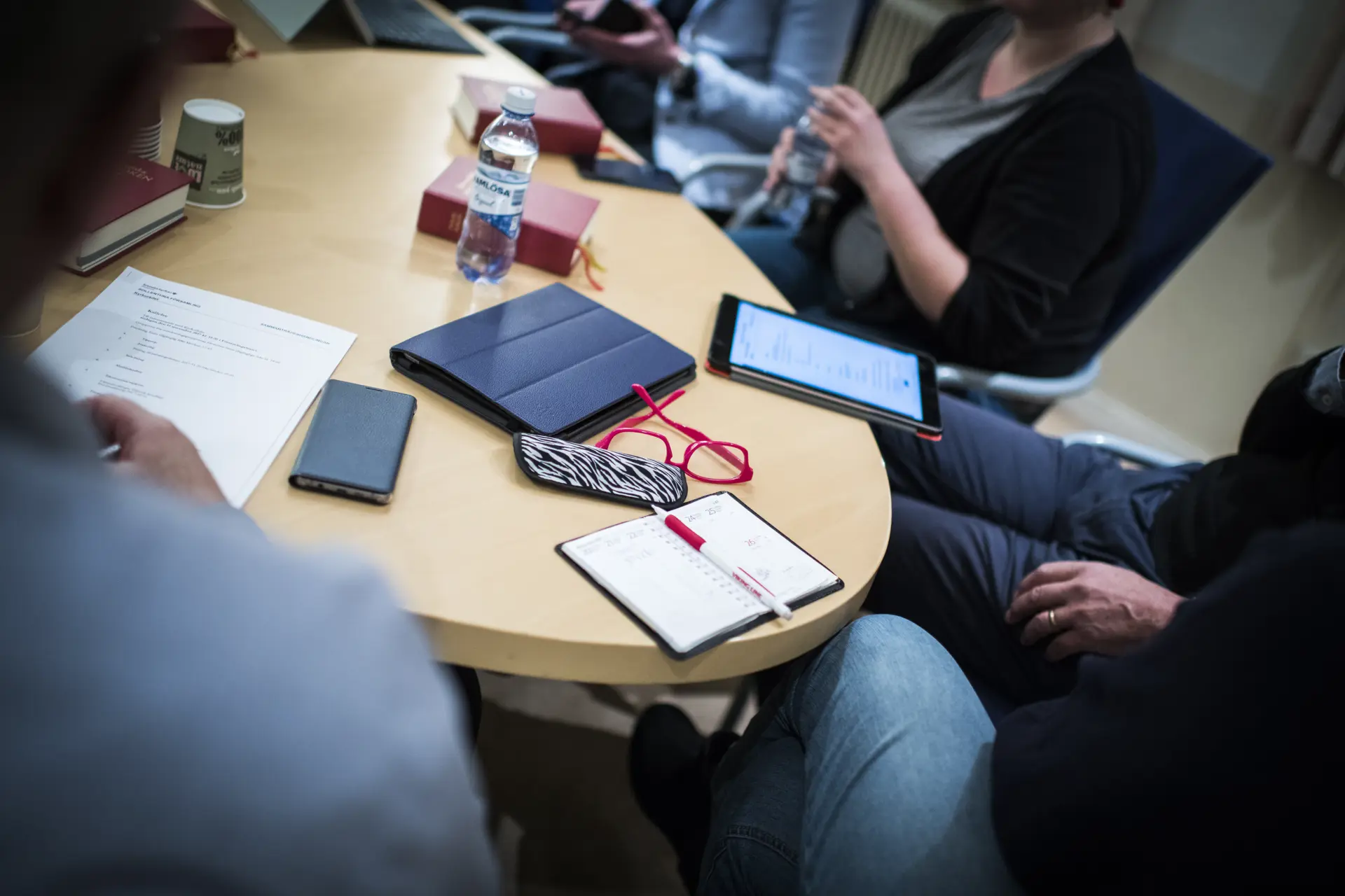 Personer runt ett bord som är fyllt av telefoner, paddor, anteckningsböcker, pennor och glasögon. 