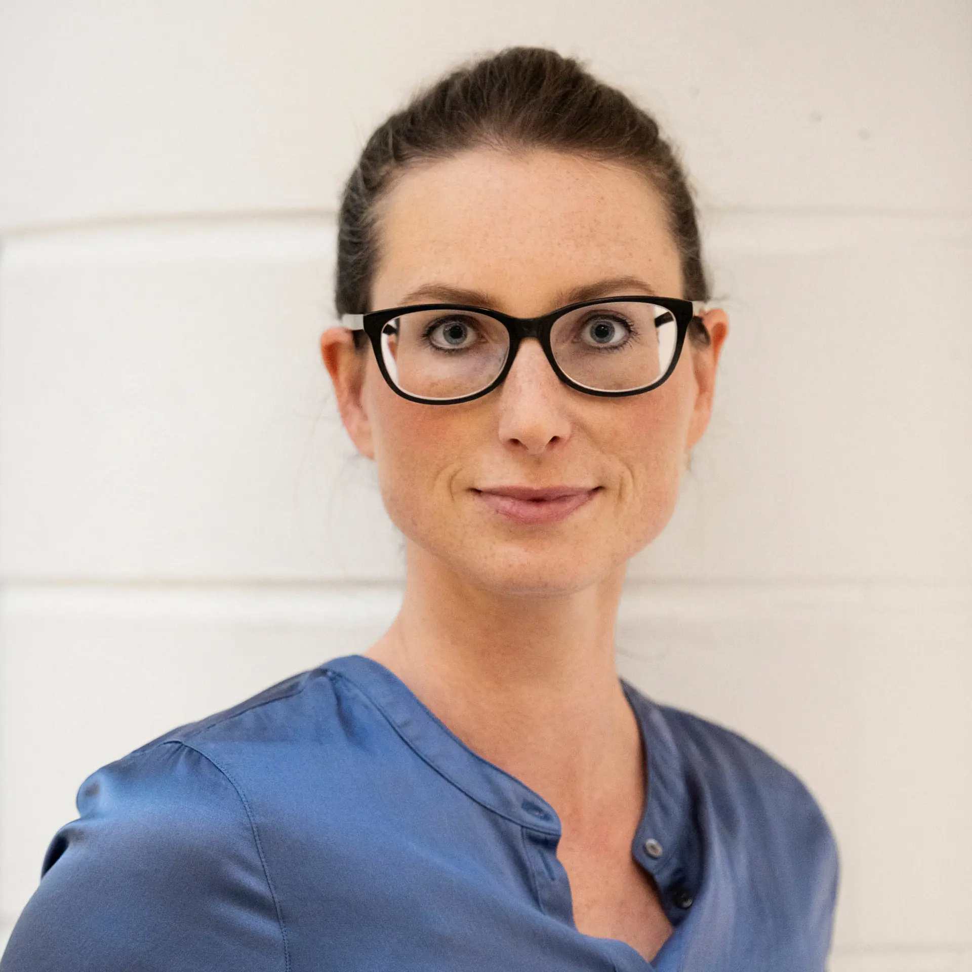 Kvinna med blå blus, bakåtkammat mörkt hår och glasögon tittar in i kameran och ler svagt. 