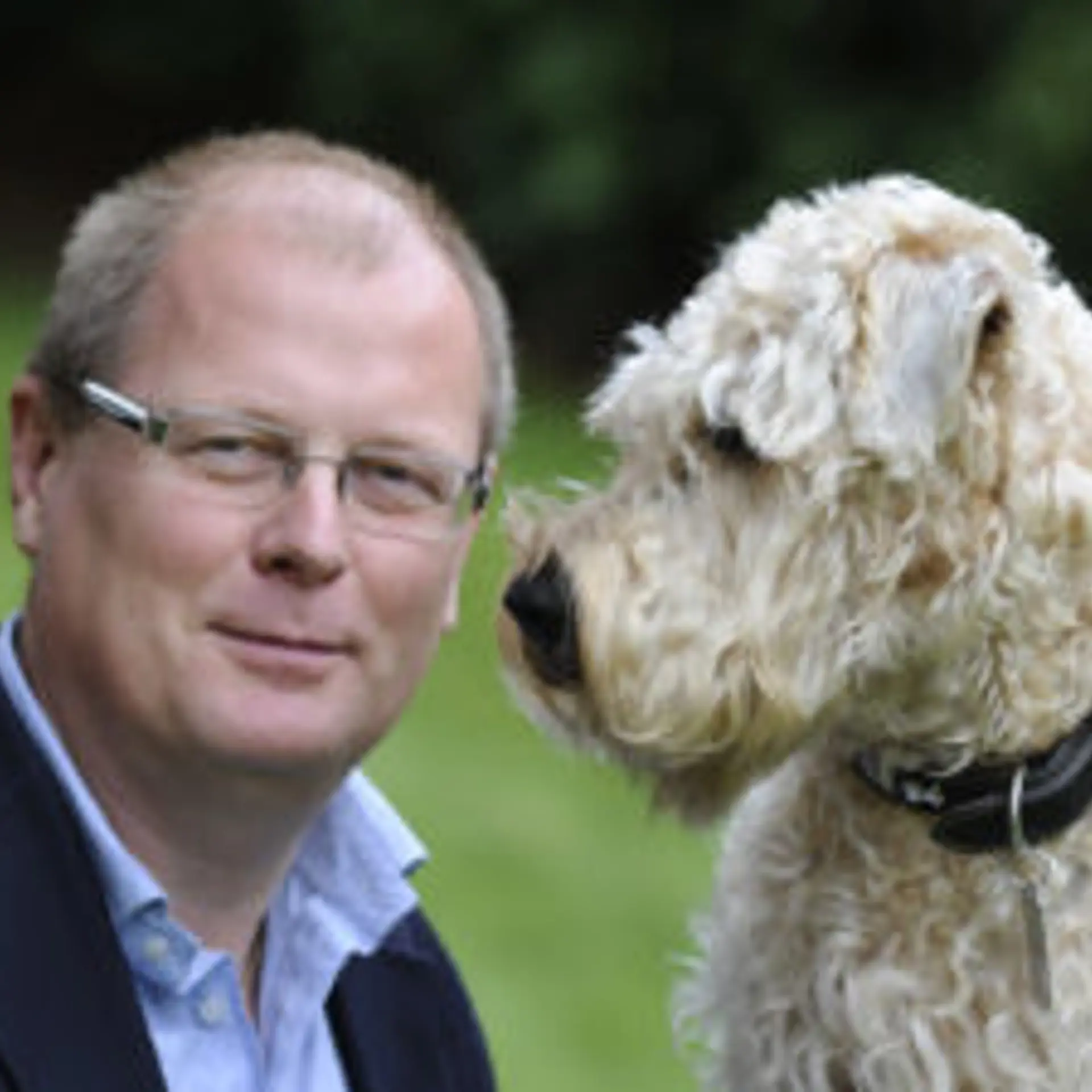 Man med blå skjorta, kavaj och glasögon tittar in i kameran. till höger i bild lurvig hund men mörkt halsband som tittar på mannen.