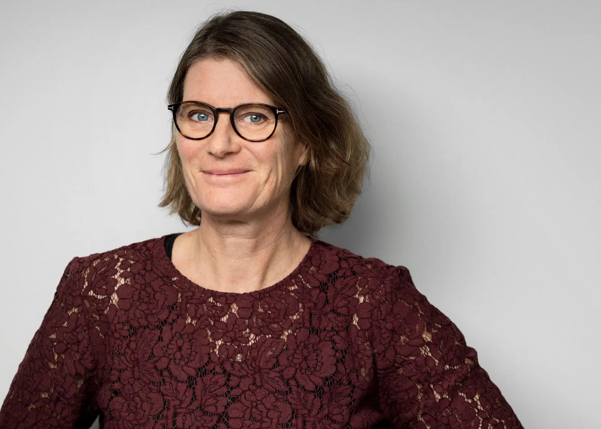 Kvinna med mörkt hår, glasögon och mörk spetstopp tittar in i kameran och ler. 