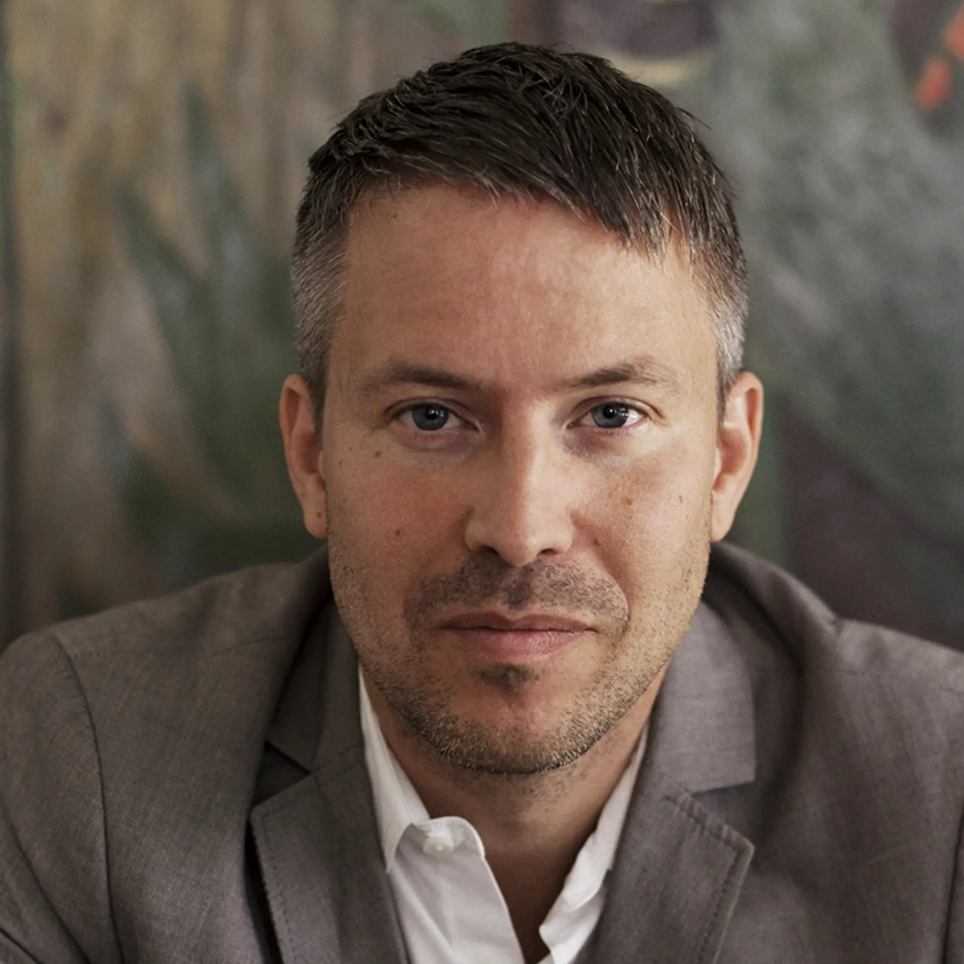 Man med mörkt hår och kort skägg, kavaj och ljus skjorta lutar sig framåt och tittar in i kameran.  