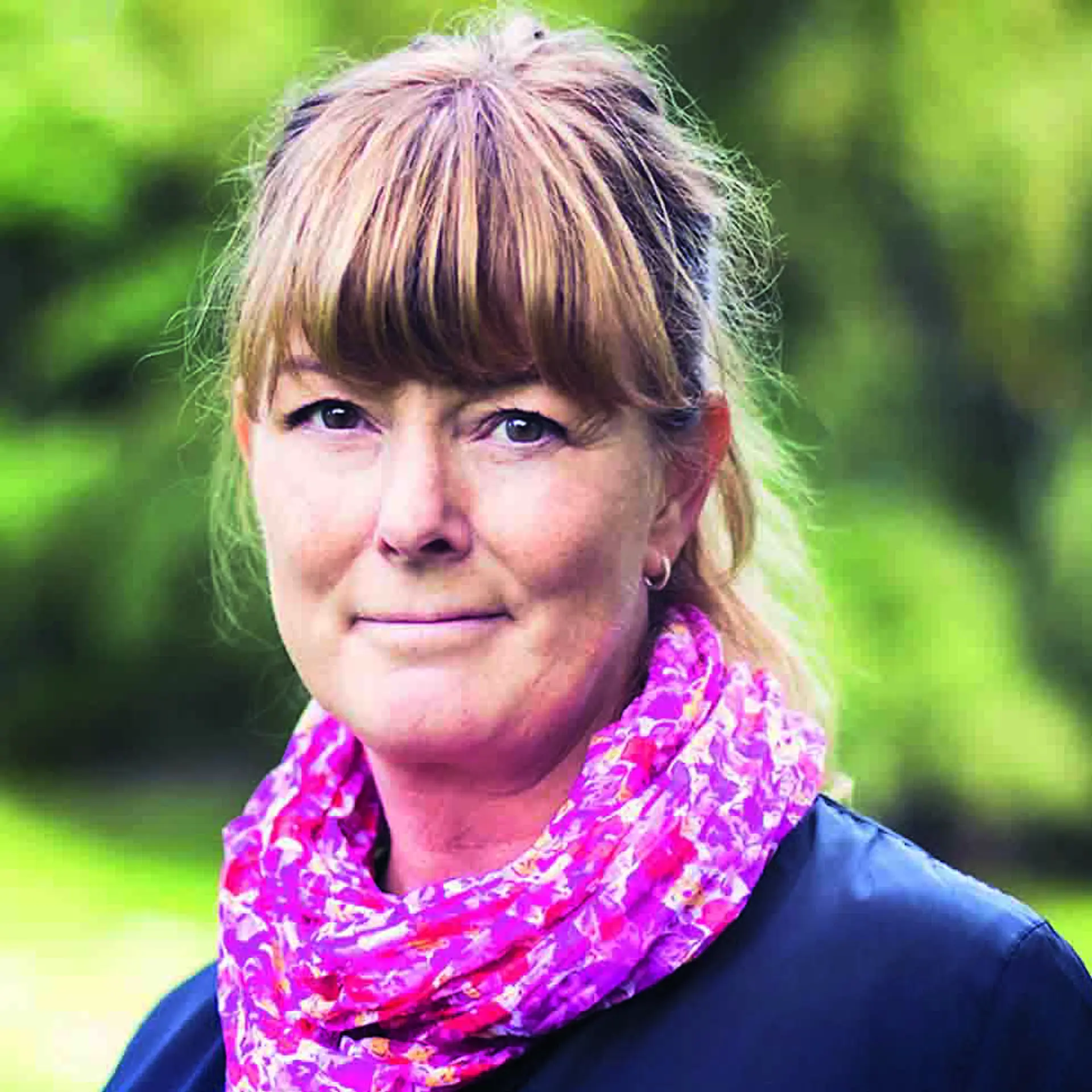 kvinna med lugg och rosamönstrad skarf runt halsen tittar in i kameran och ler svagt. 