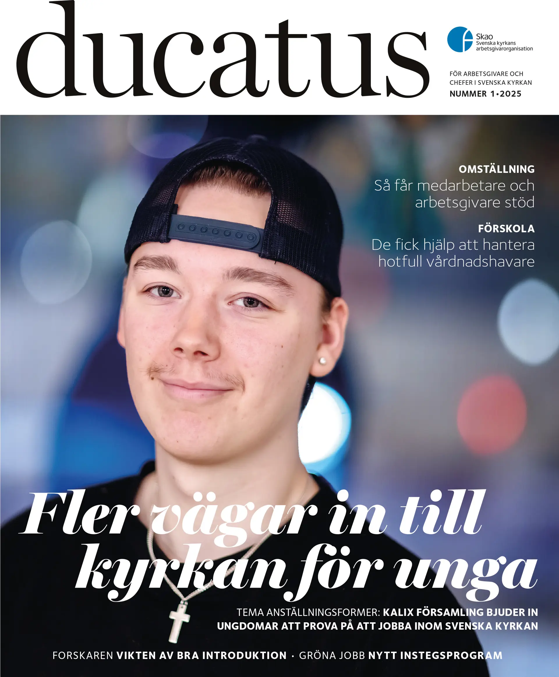 En ung pojke med bak och framvänd svart keps, mörk tröja och smycke i form av ett kors runt halsen tittar in i kameran och ler. 