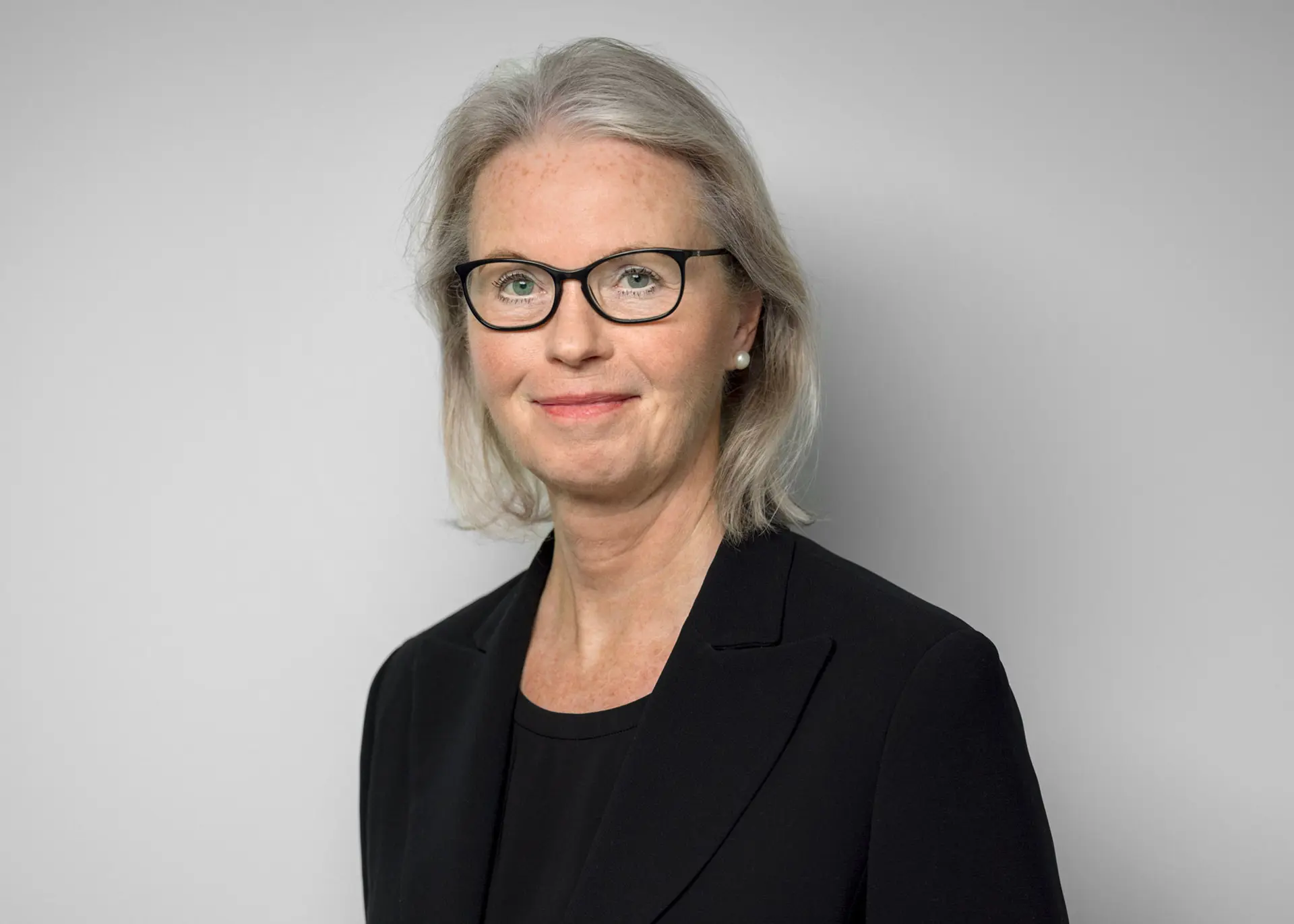 Kvinna med mörka kläder, mörka glasögon och litet smycke runt halsen tittar in i kameran och ler.