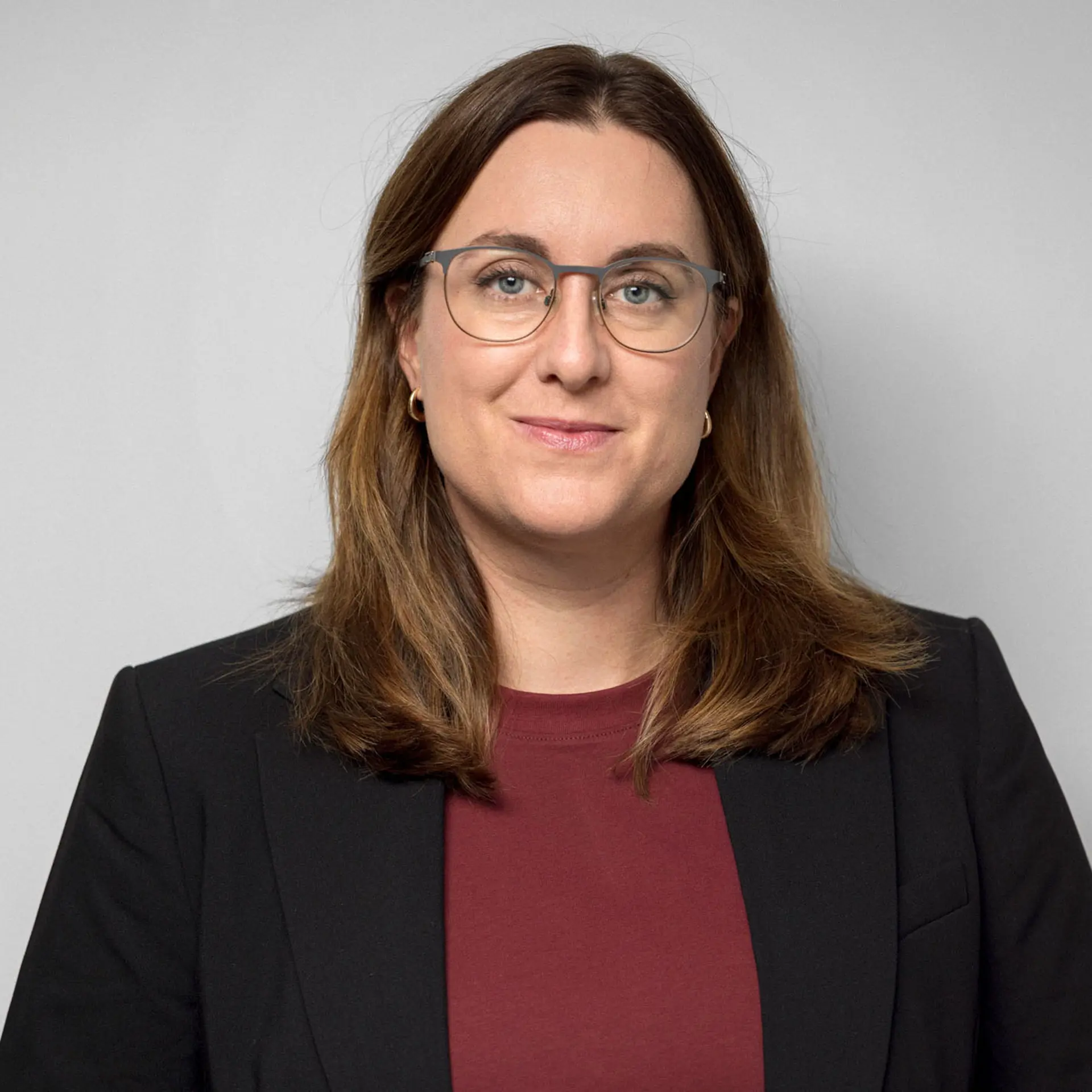 Kvinna med purpurfärgad topp under mörk kavaj, axellångt hår och glasögon. Tittar in i kameran och ler svagt. 