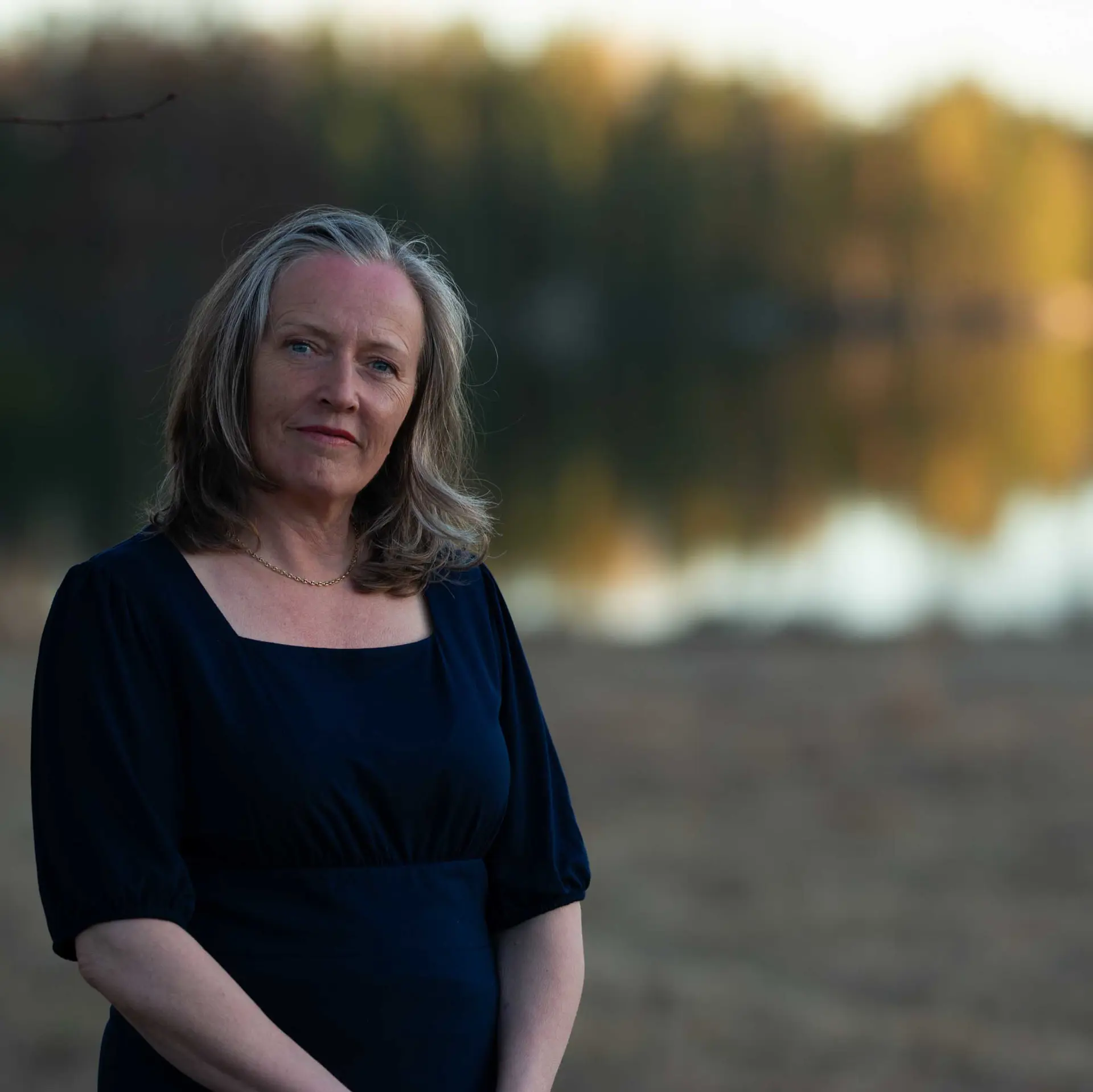 Kvinna med mörkt axellångt hår och mörk tröja står intill vatten, och ser in i kameran. 