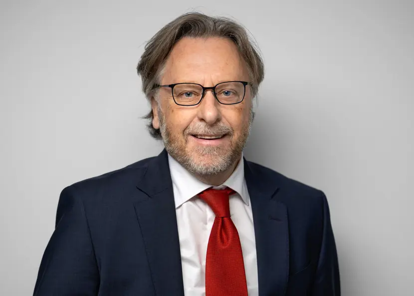 Man med vit skjorta, röd slips och mörk kavaj, kort skägg och mörka glasögon tittar in i kameran och ler. 