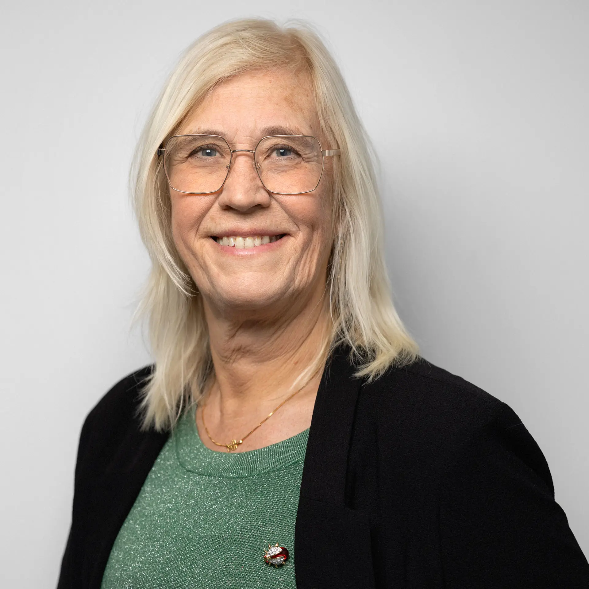 Kvinna med blont axellångt hår, glasögon, grön tröja och svart kavaj tittar in i kameran och ler.