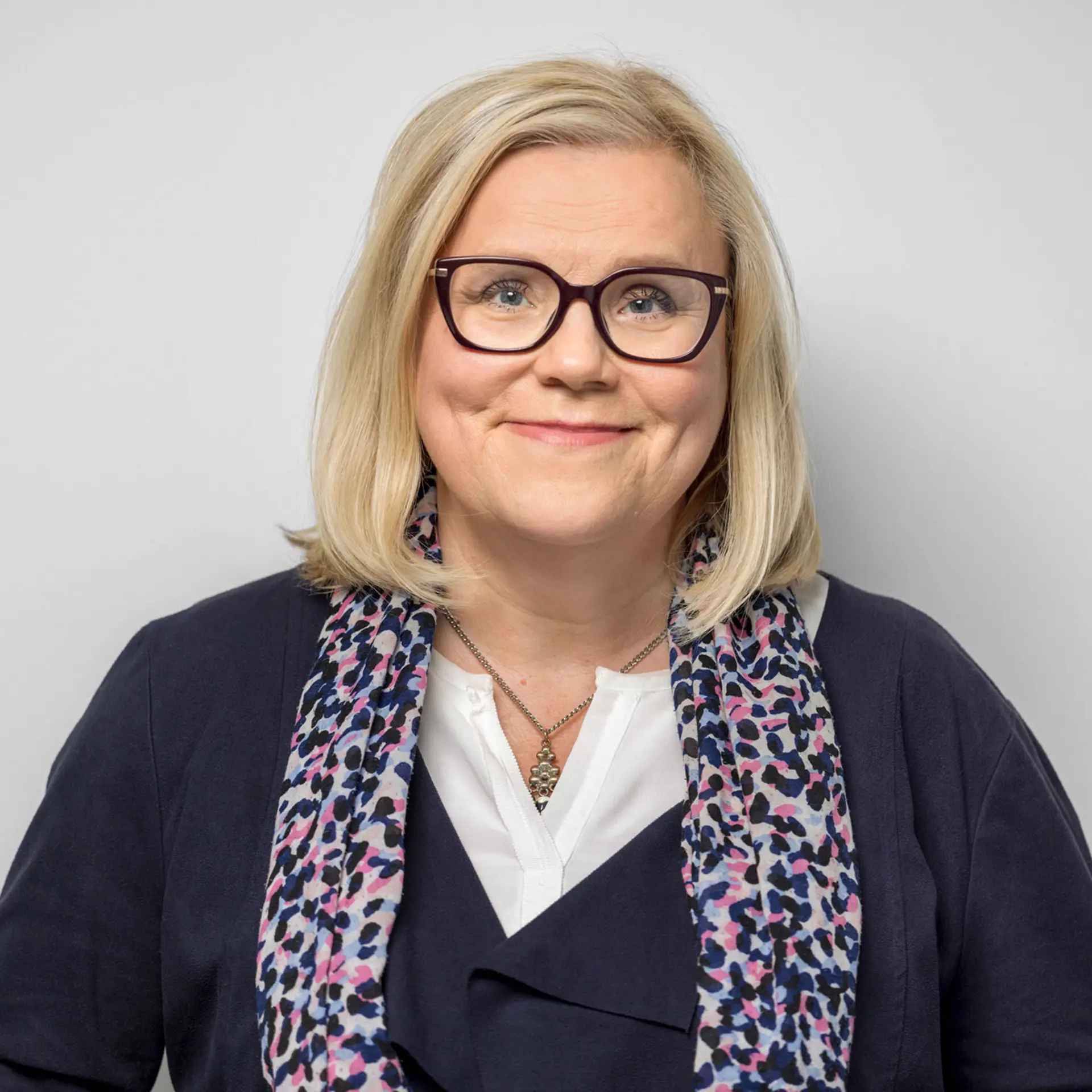 Kvinna med blont axellångt hår, vit blus, glasögon, mörk kavaj tittar in i kameran och ler.. 